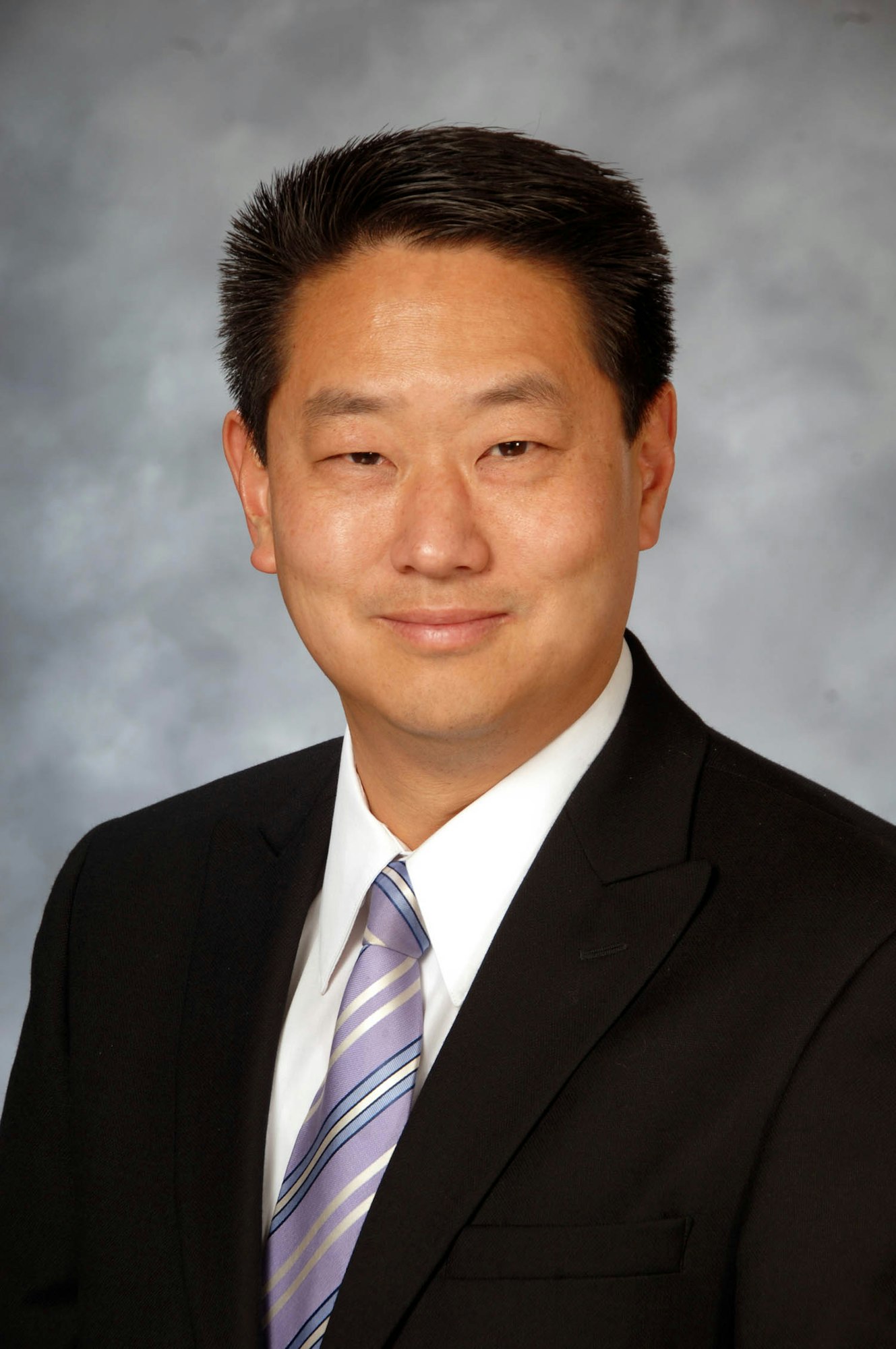 A person wearing a black suit and a striped tie against a gray background.