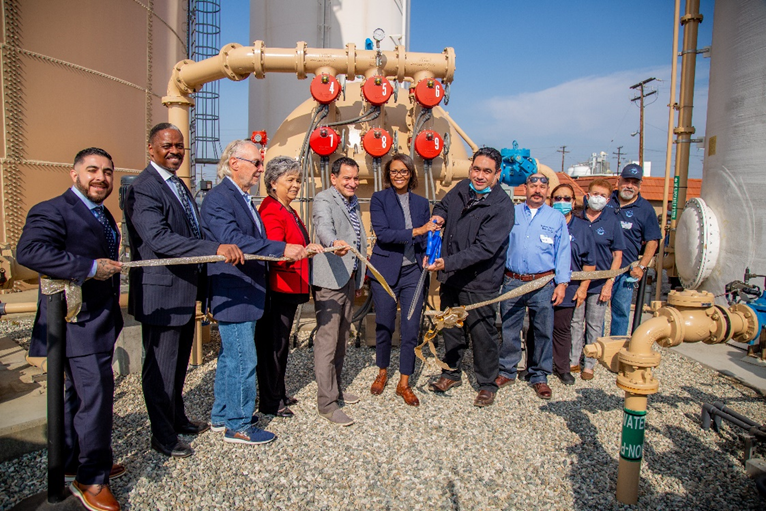 A group of people at a ribbon-cutting ceremony near industrial equipment, celebrating a new facility or project.