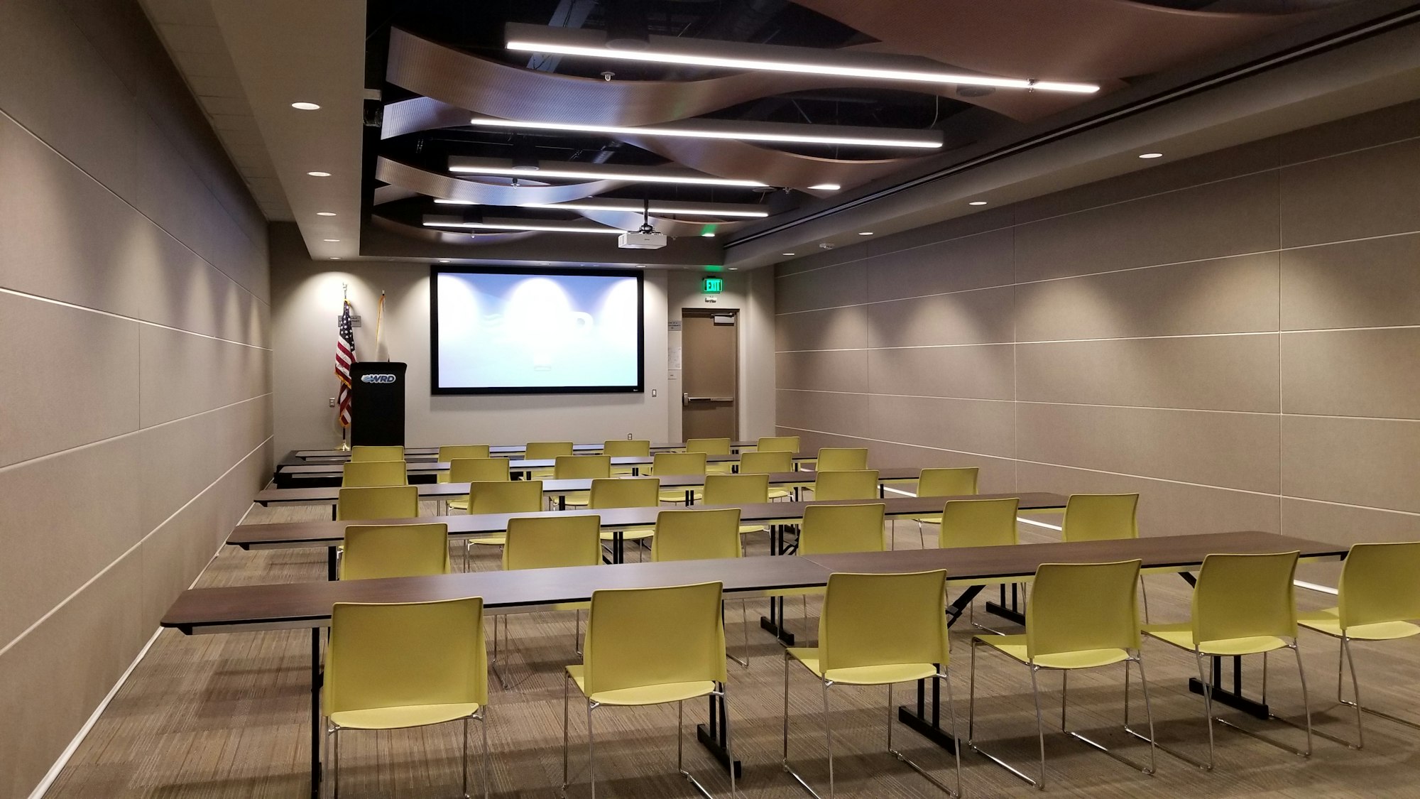 A lecture room with yellow chairs, tables, a projector screen, podium, and an American flag.
