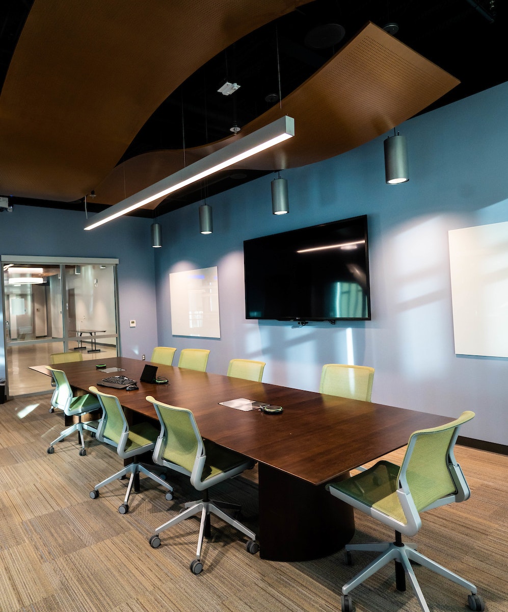 Modern conference room with a long table, green chairs, TV screen on the wall, and large lighting fixtures above.