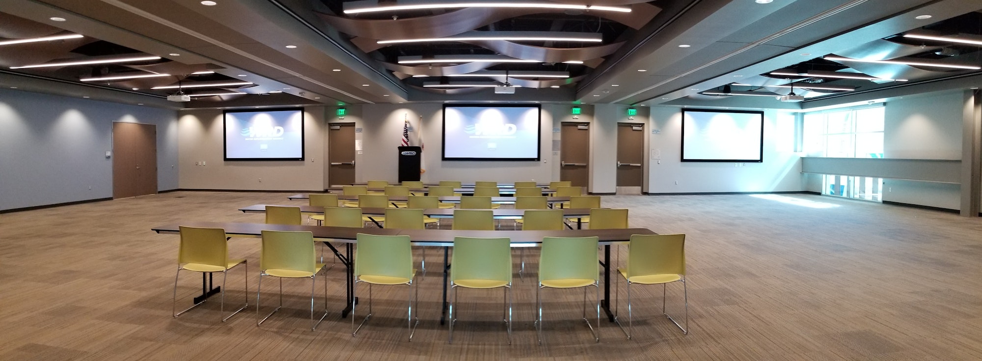 A modern conference room with yellow chairs, tables, two large screens, and a podium.