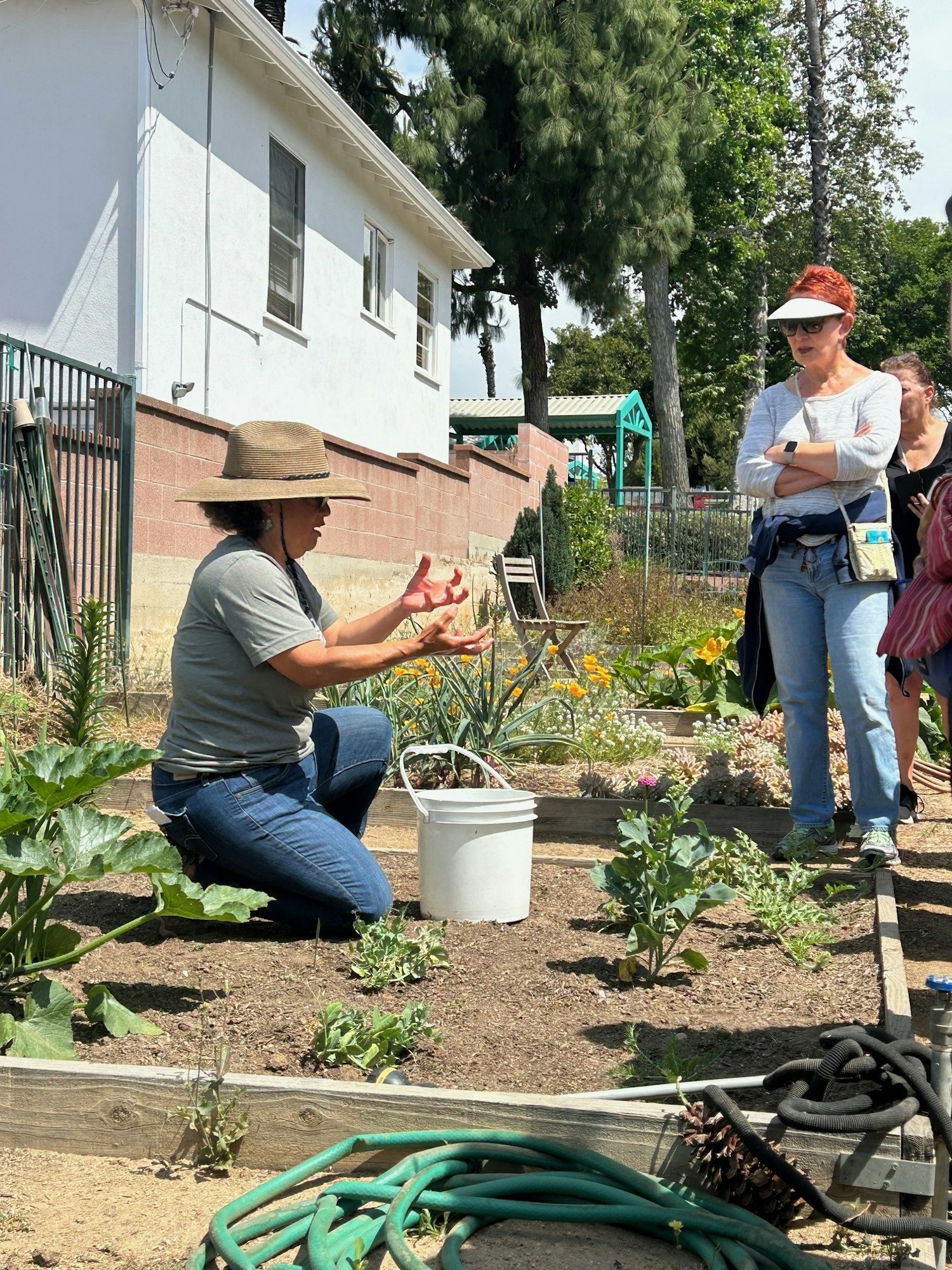 A woman in a hat demonstrates gardening techniques to a group in a community garden on a sunny day.