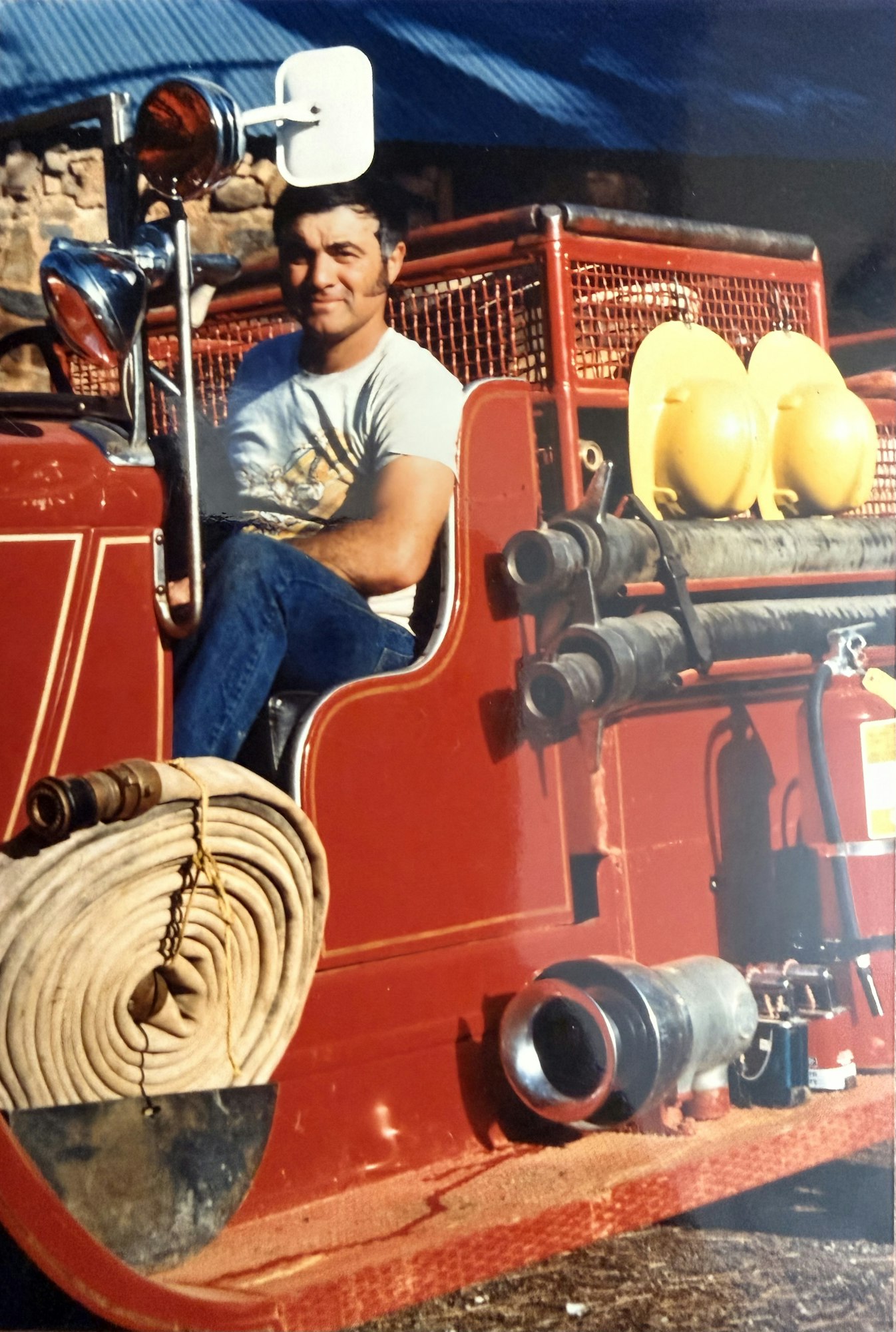 A man sits on a red fire truck, smiling, with fire hoses and equipment visible in the background.