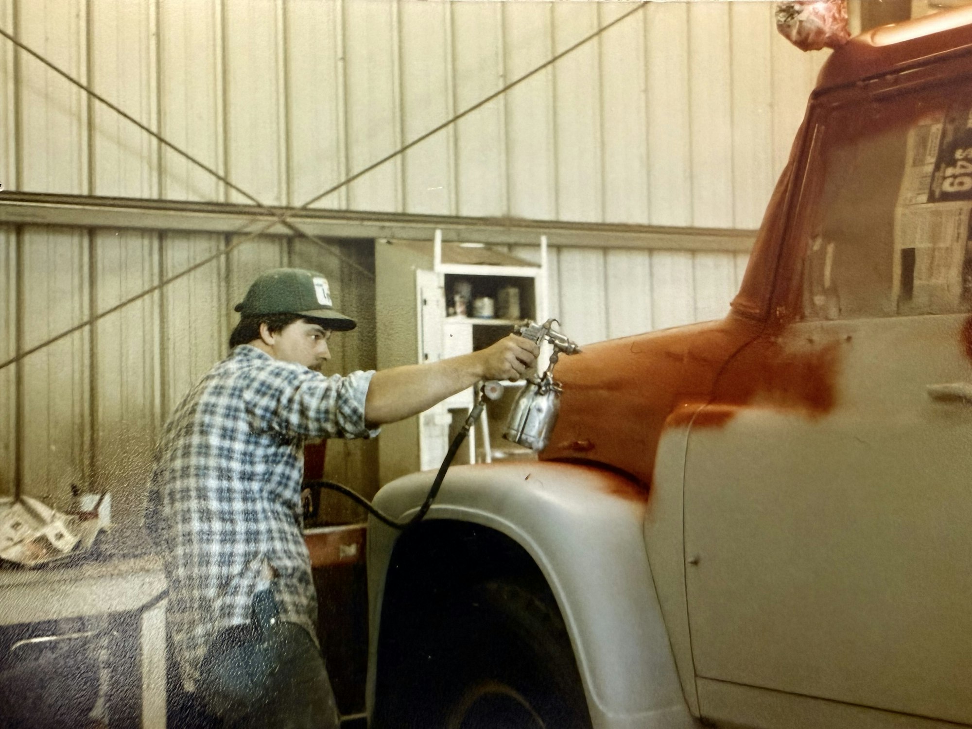 A person is spray painting a truck's fender in a workshop, wearing a plaid shirt and cap, focused on the task.
