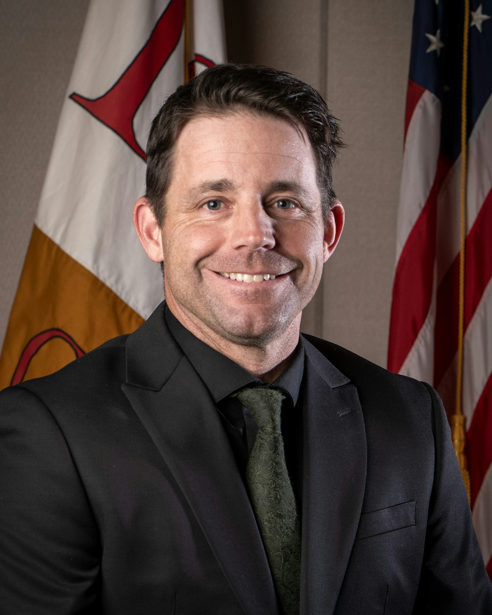 A man in a suit with a green tie smiles in front of two flags.