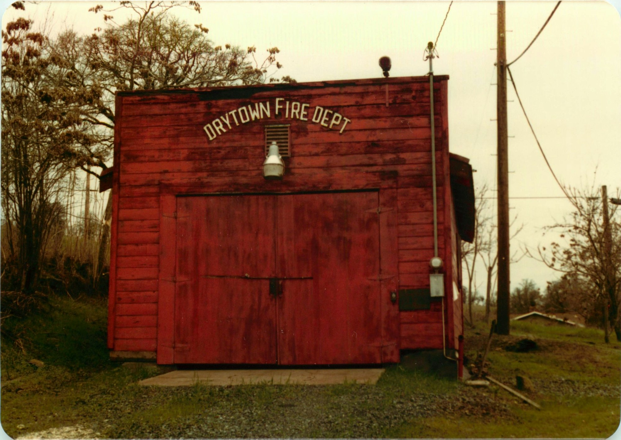 A rustic red wooden building labeled "Drytown Fire Dept," surrounded by trees and power lines, set in a rural area.