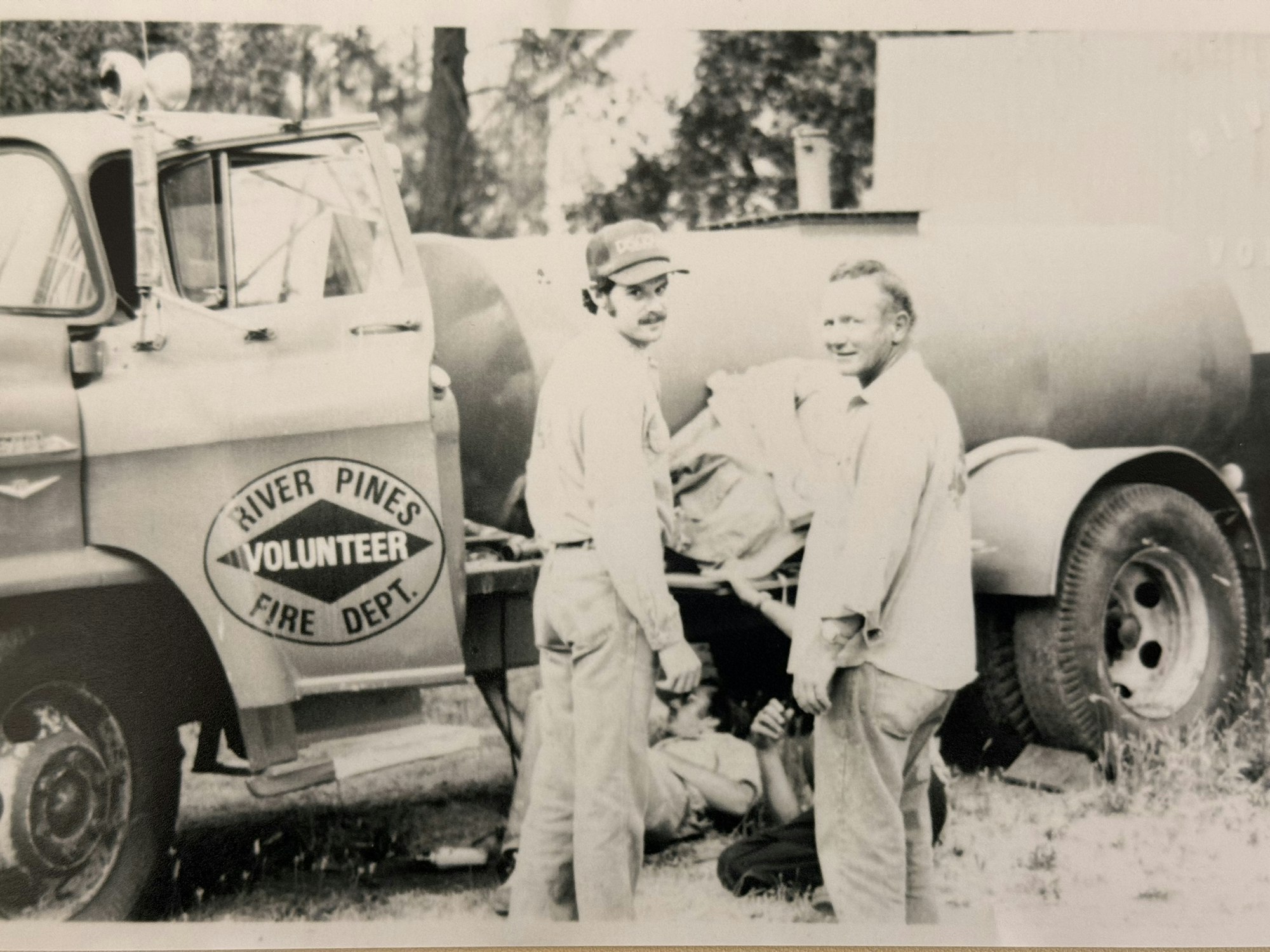 The image shows two men beside a vintage fire truck marked "River Pines Volunteer Fire Dept," suggesting a historic firefighting context.