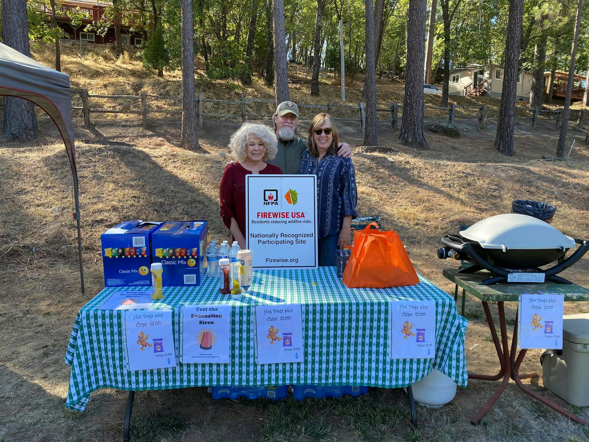 Three people at an outdoor booth with "Firewise USA" sign, snacks, and a grill.