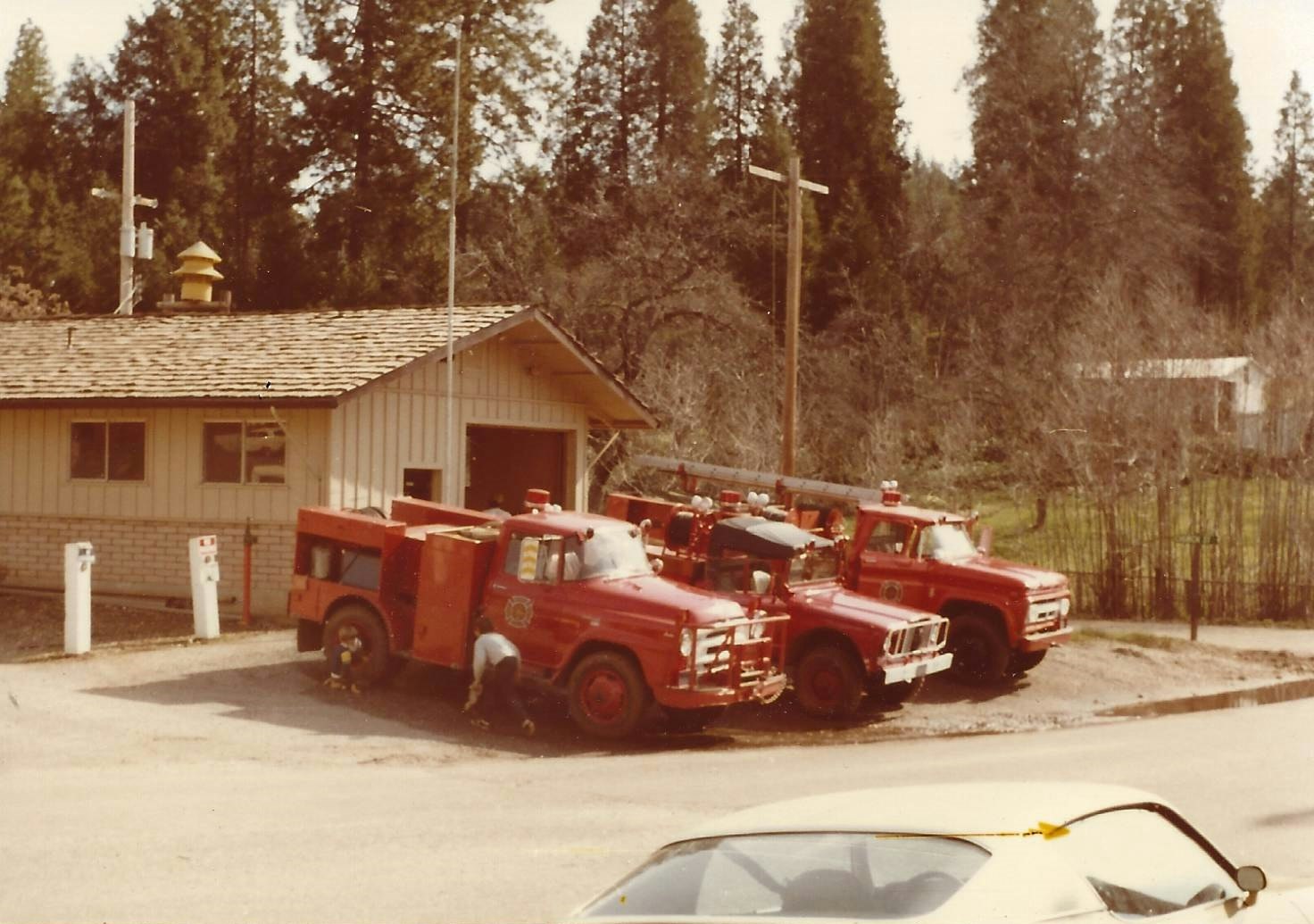 The image shows two red fire trucks near a building, with a person working on one and trees in the background.