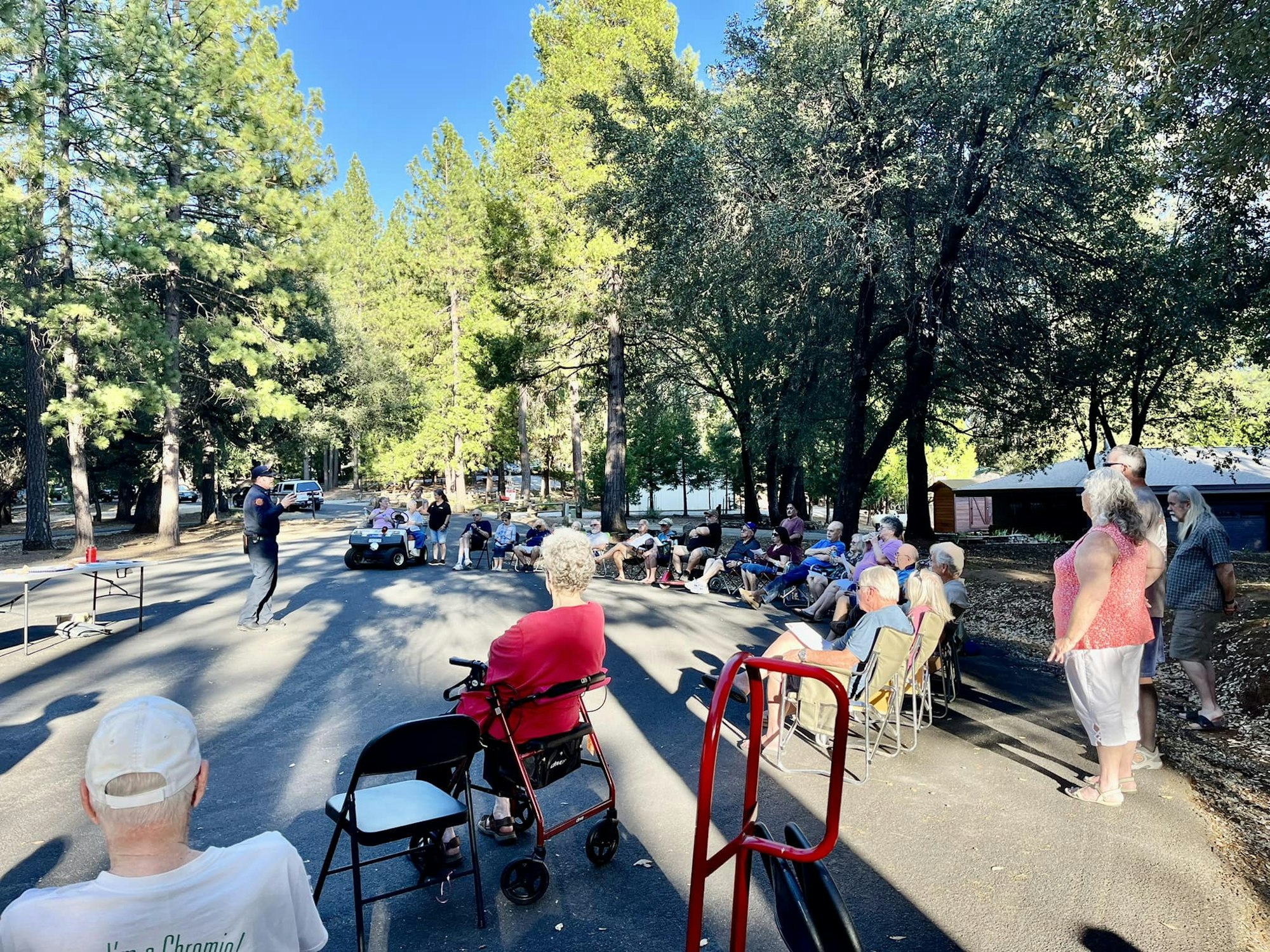A group of people outdoors, seated and standing on a road surrounded by trees, listening to a person speaking.