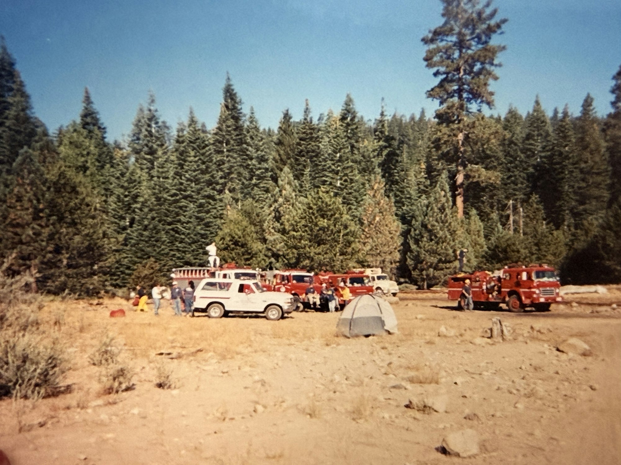 A gathering in a forested area with fire trucks, people, and a tent, likely for a community or emergency response event.