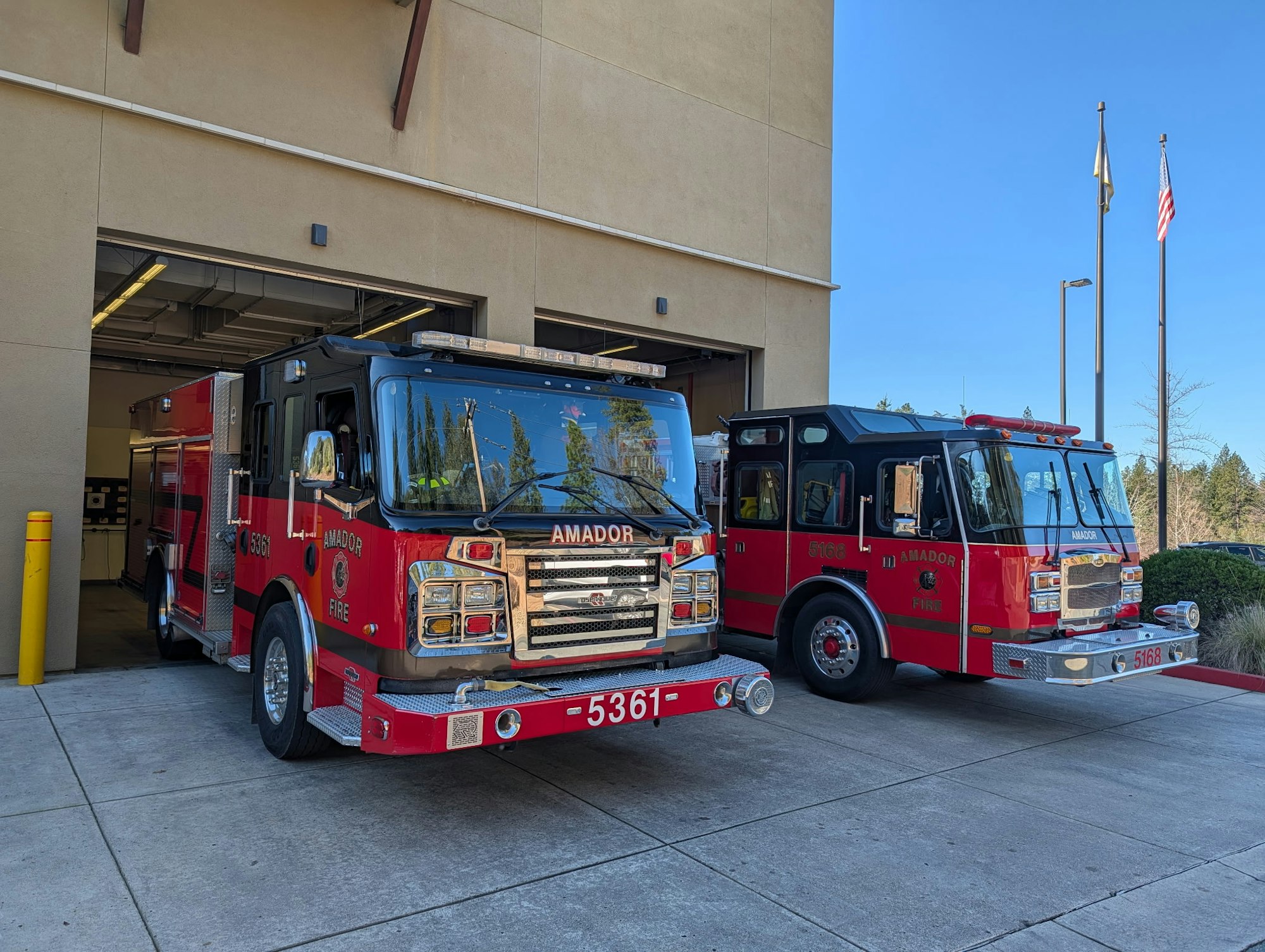 Two red fire trucks parked outside a fire station, with flags in the background under a clear blue sky.