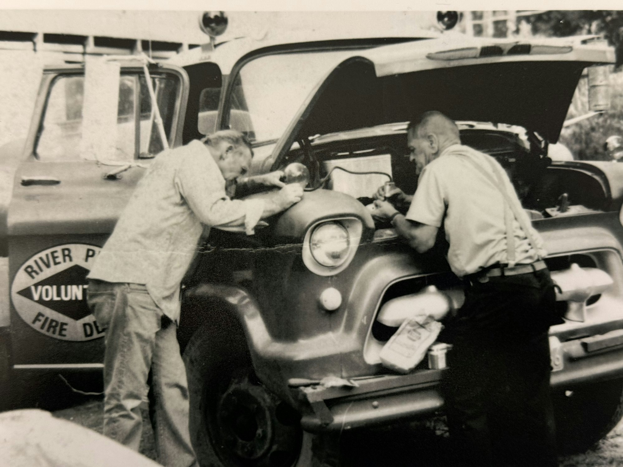 Two men are working on the engine of an old fire truck, focusing on repairs in a black-and-white photo.