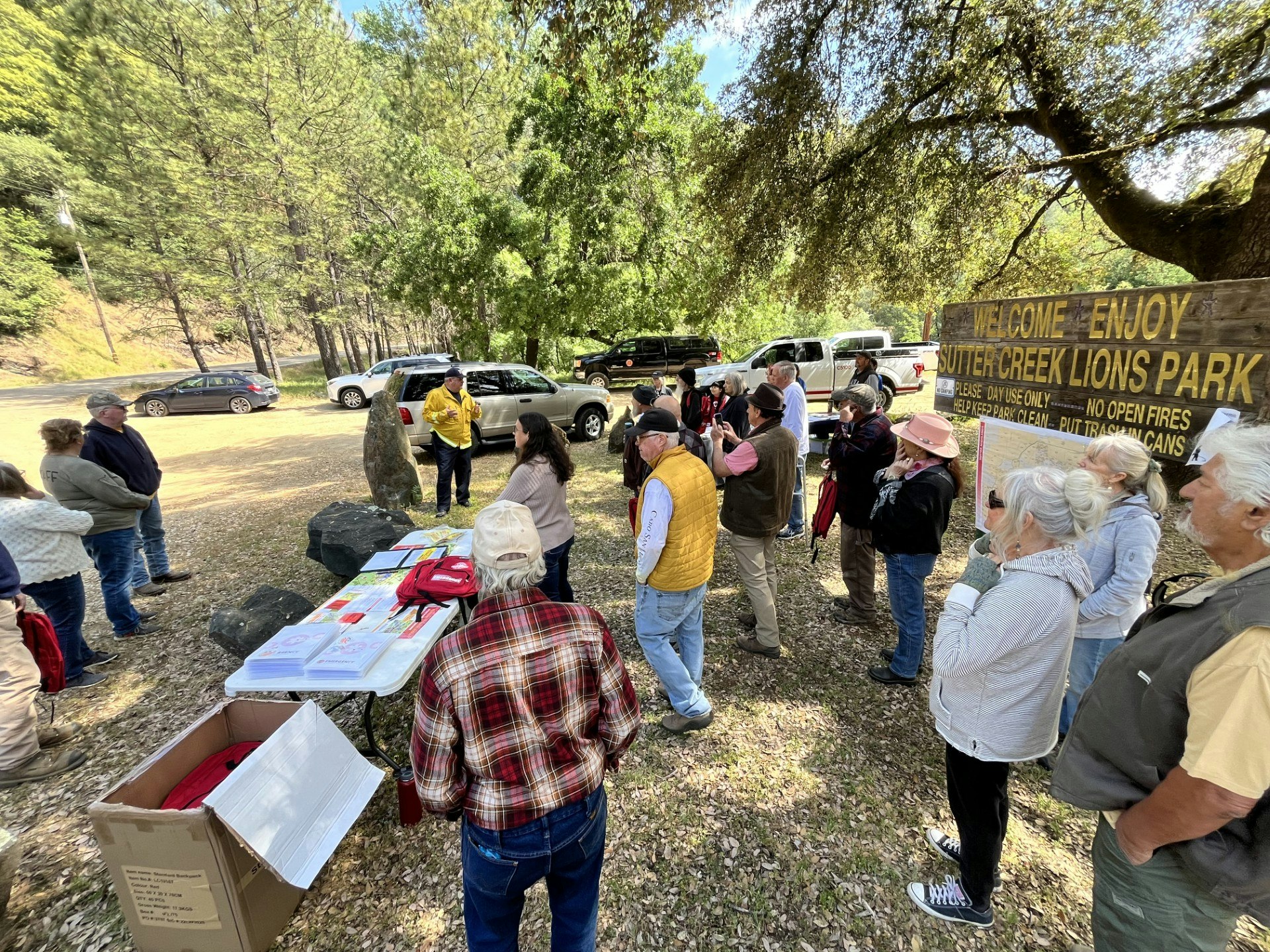 A group of people gathered outdoors at Sutter Creek Lions Park, with a table of materials and parked cars nearby.