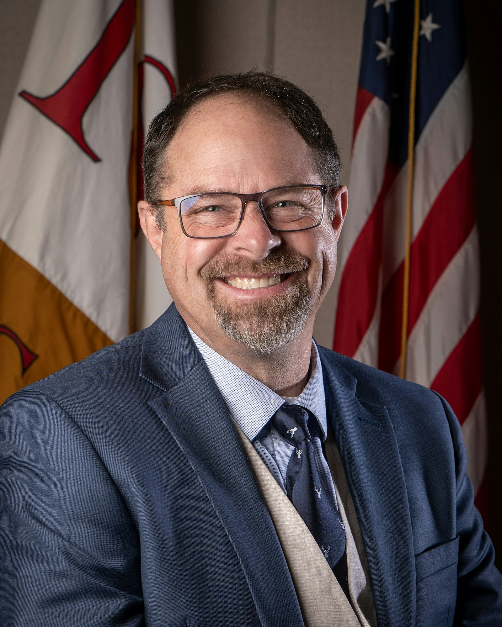 A smiling man in a suit with glasses stands in front of two flags, including the American flag.