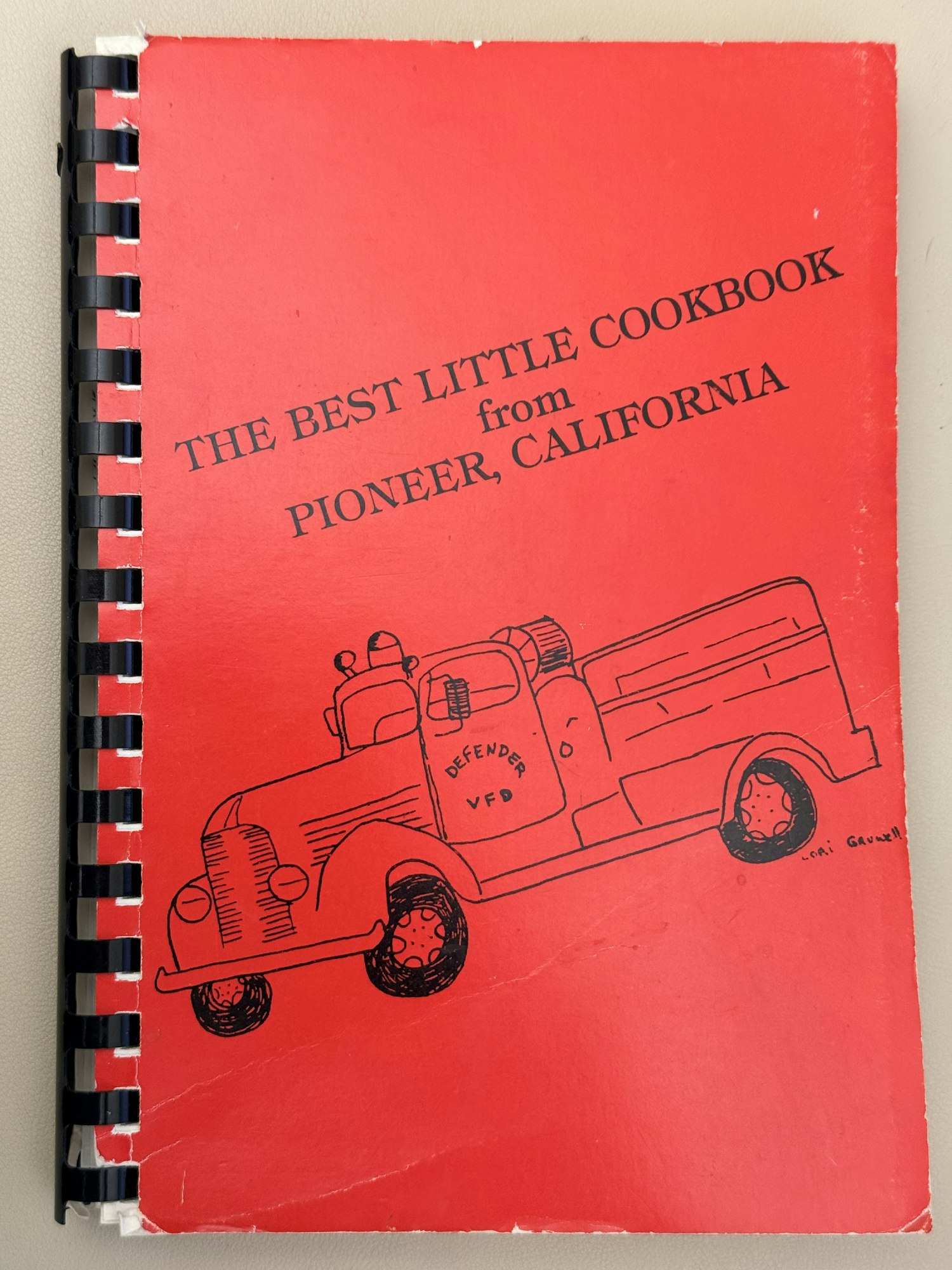 A red cookbook titled "The Best Little Cookbook from Pioneer, California," featuring an illustration of an old-fashioned vehicle.