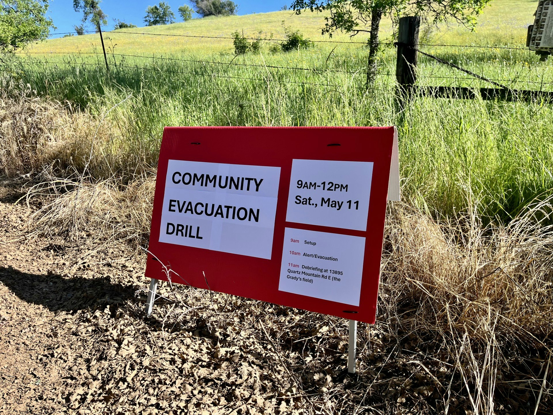 Red sign for a community evacuation drill on Saturday, May 11, 9AM-12PM, with a schedule in a grassy area.
