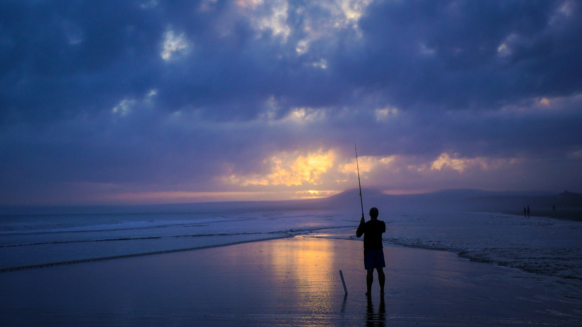 Silhouette of a person fishing on a beach at sunset under a dramatic, cloudy sky.