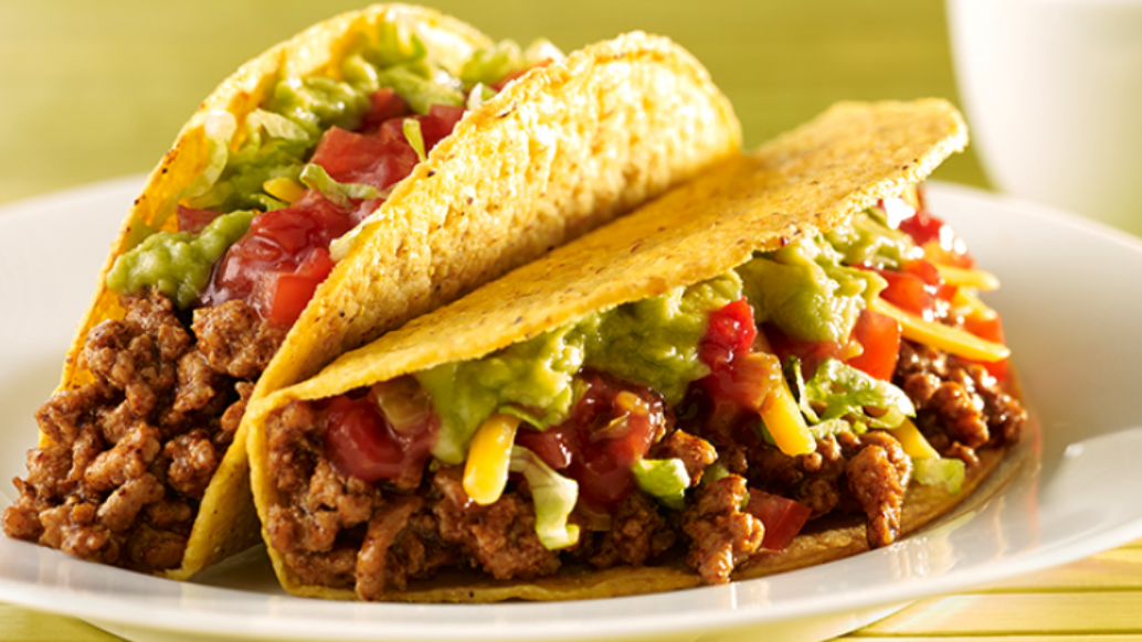 Two tacos with ground beef, guacamole, tomatoes, lettuce, and cheese in hard taco shells on a white plate.