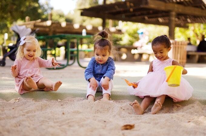 Three children playing in a sandbox; one holds a yellow bucket. They're enjoying a sunny day outdoors.