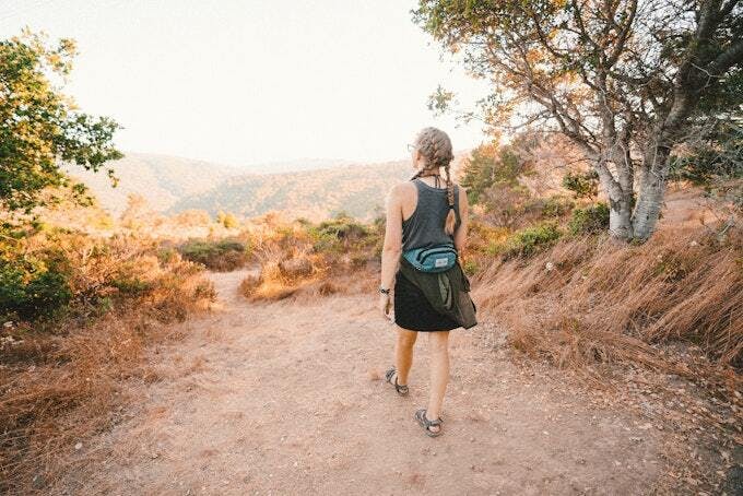 A person with a braid hikes on a dirt path through a scenic landscape, surrounded by dry grass and trees.