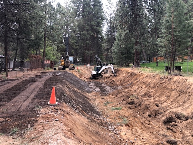 A construction site in a wooded area, featuring machinery, a dirt trench, and an orange traffic cone.