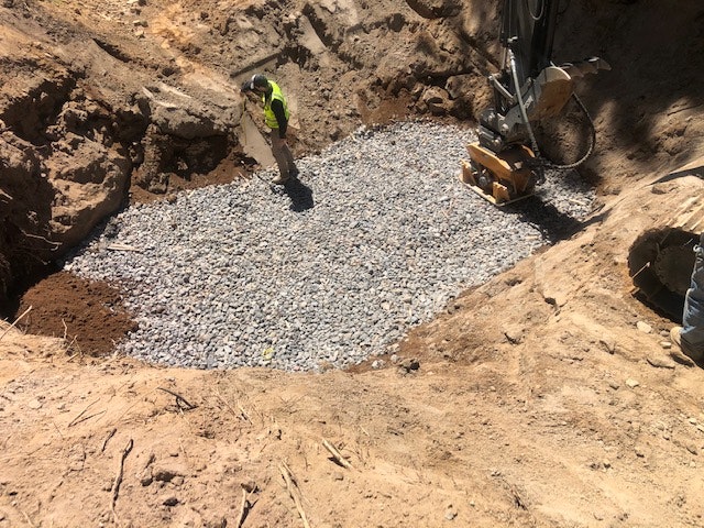 A construction site shows a worker in a pit filled with gravel, surrounded by earth and machinery.