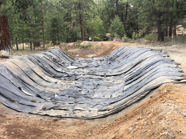 The image shows a wrinkled black tarp or liner spread over a dirt area, surrounded by trees in a natural setting.