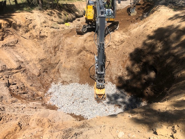 An excavator is digging a site, revealing gravel and dirt in a construction area surrounded by trees.