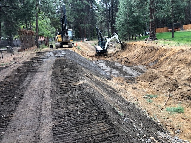 The image shows construction equipment working on a dirt pathway, with one machine digging and another near the edge of a trench.