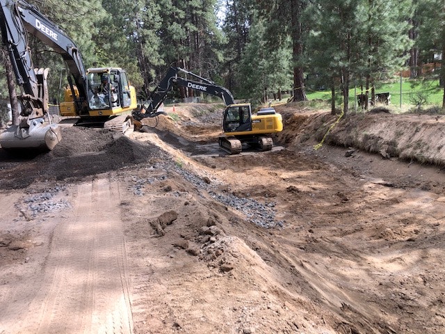 Construction site with two excavators working on a dirt trench surrounded by trees.