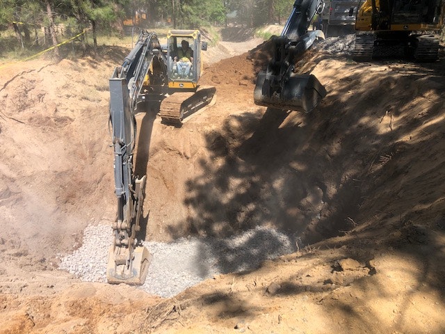 Excavators working in a dusty trench, placing gravel, surrounded by trees and earth-moving equipment.