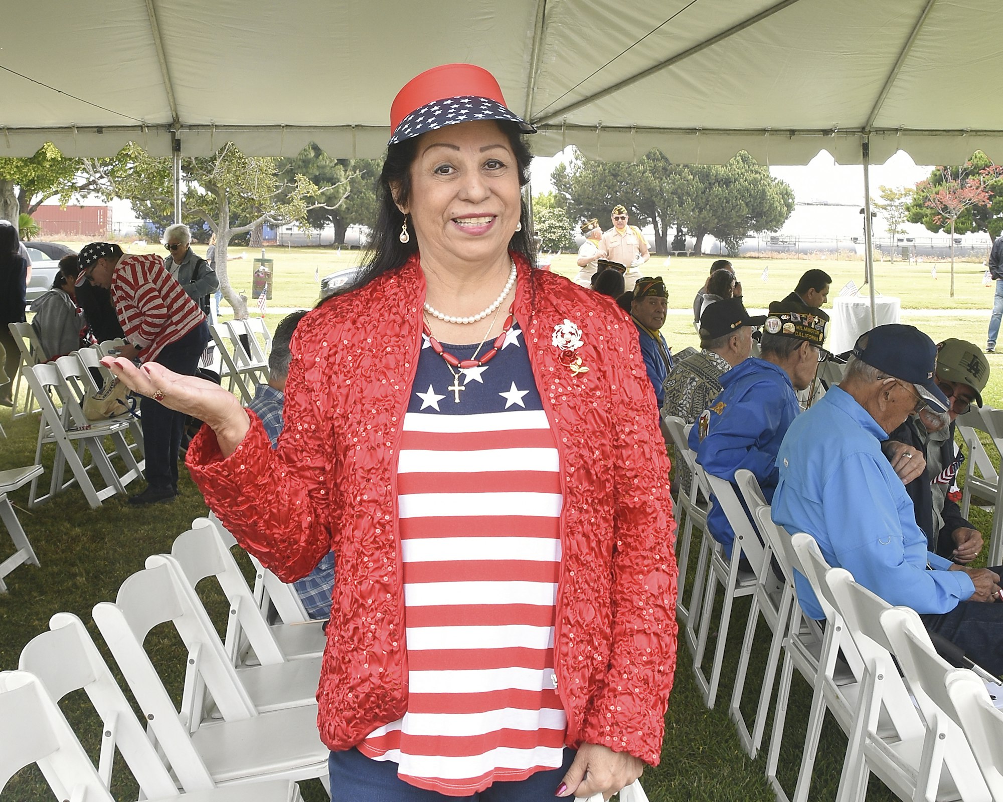 A woman in patriotic attire stands smiling under a tent with seated people, possibly at an outdoor event or gathering.