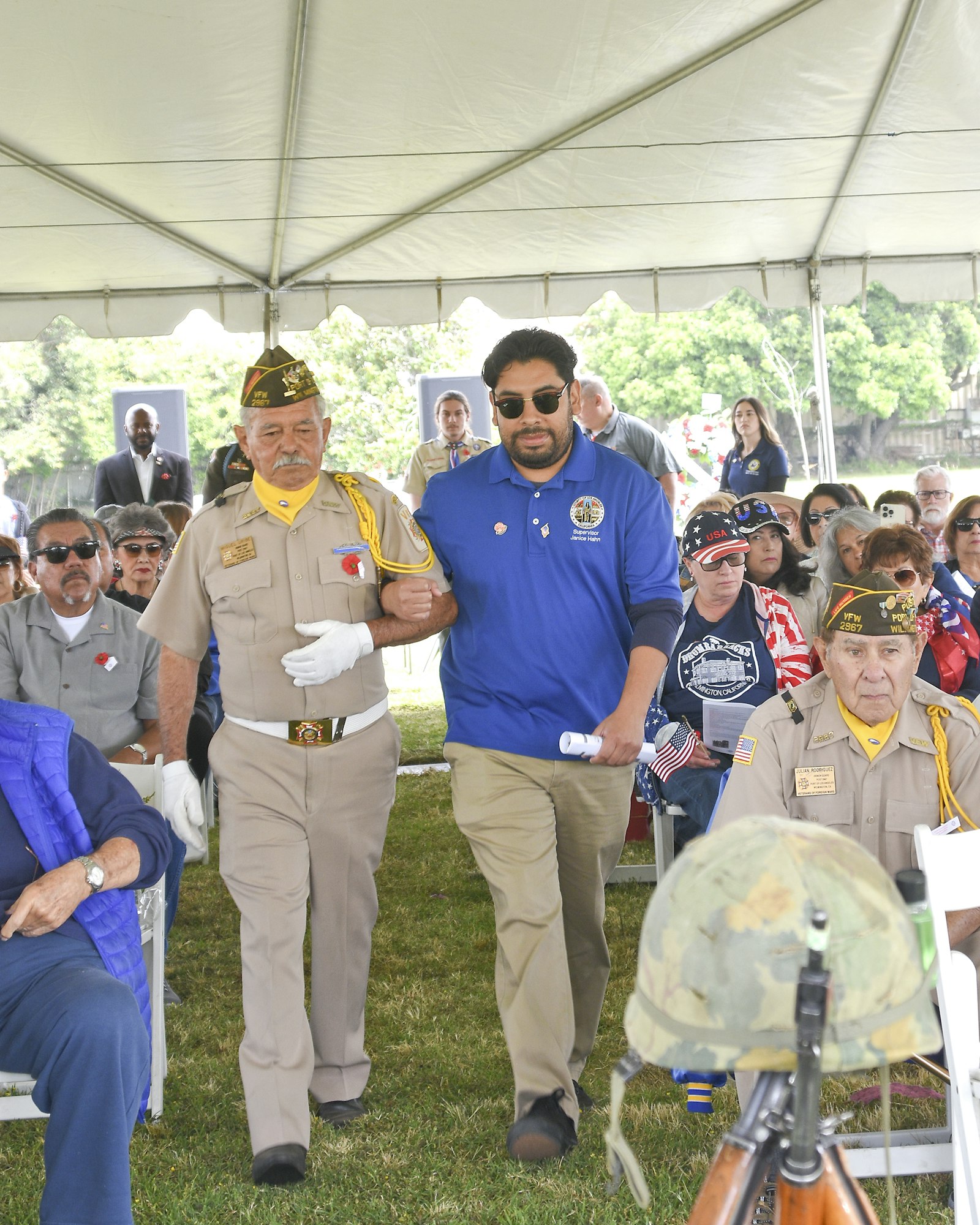 People at an outdoor event, possibly a military or veteran ceremony, under a tent with an audience.
