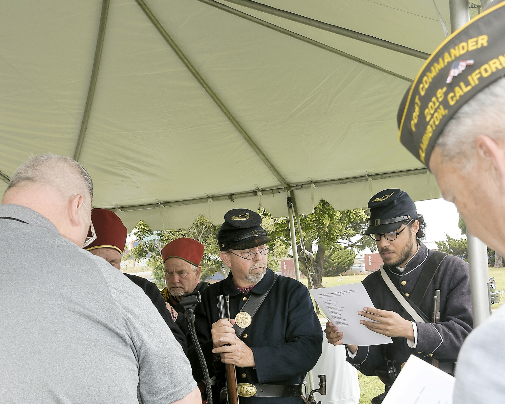 Group of people in historical military uniforms under a tent, one reading a document.