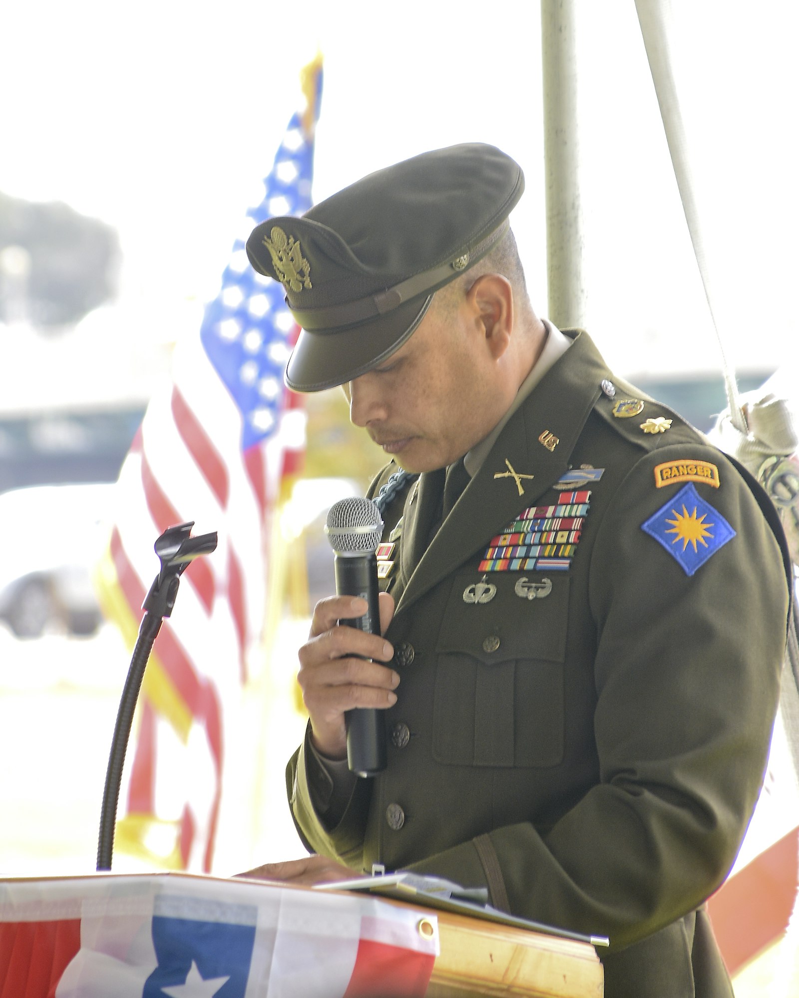 A uniformed military officer speaking into a microphone with U.S. flags in the background.