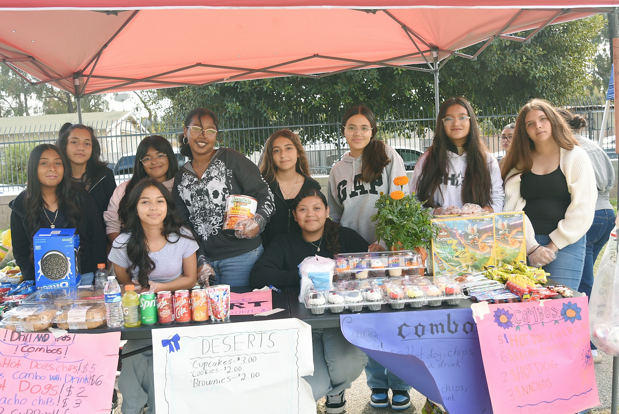 Group of people at a food stand with snacks and handwritten signs displaying prices for various items.