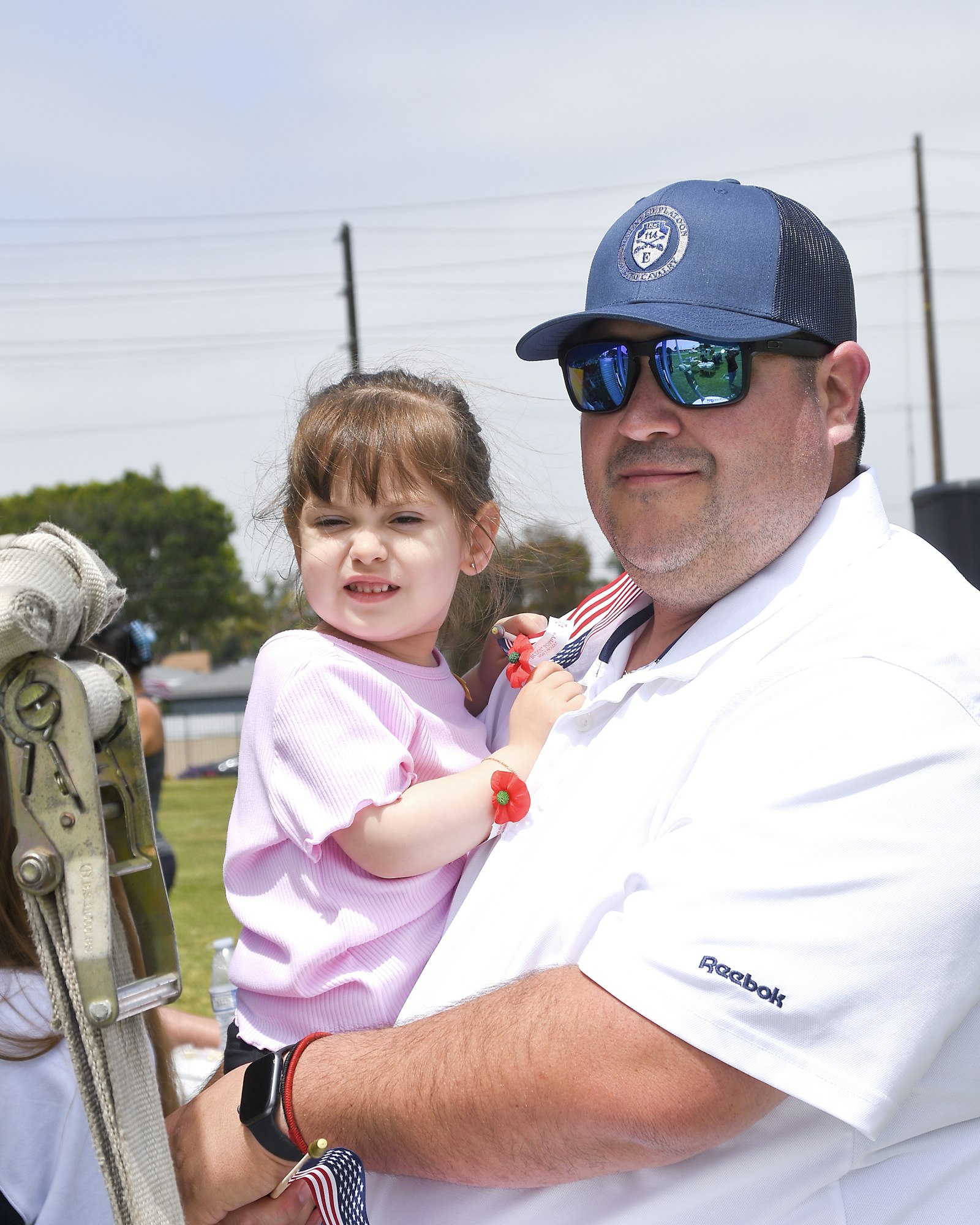 A man in sunglasses and cap carrying a child next to a rope barrier, both looking to the side, in a bright outdoor setting.