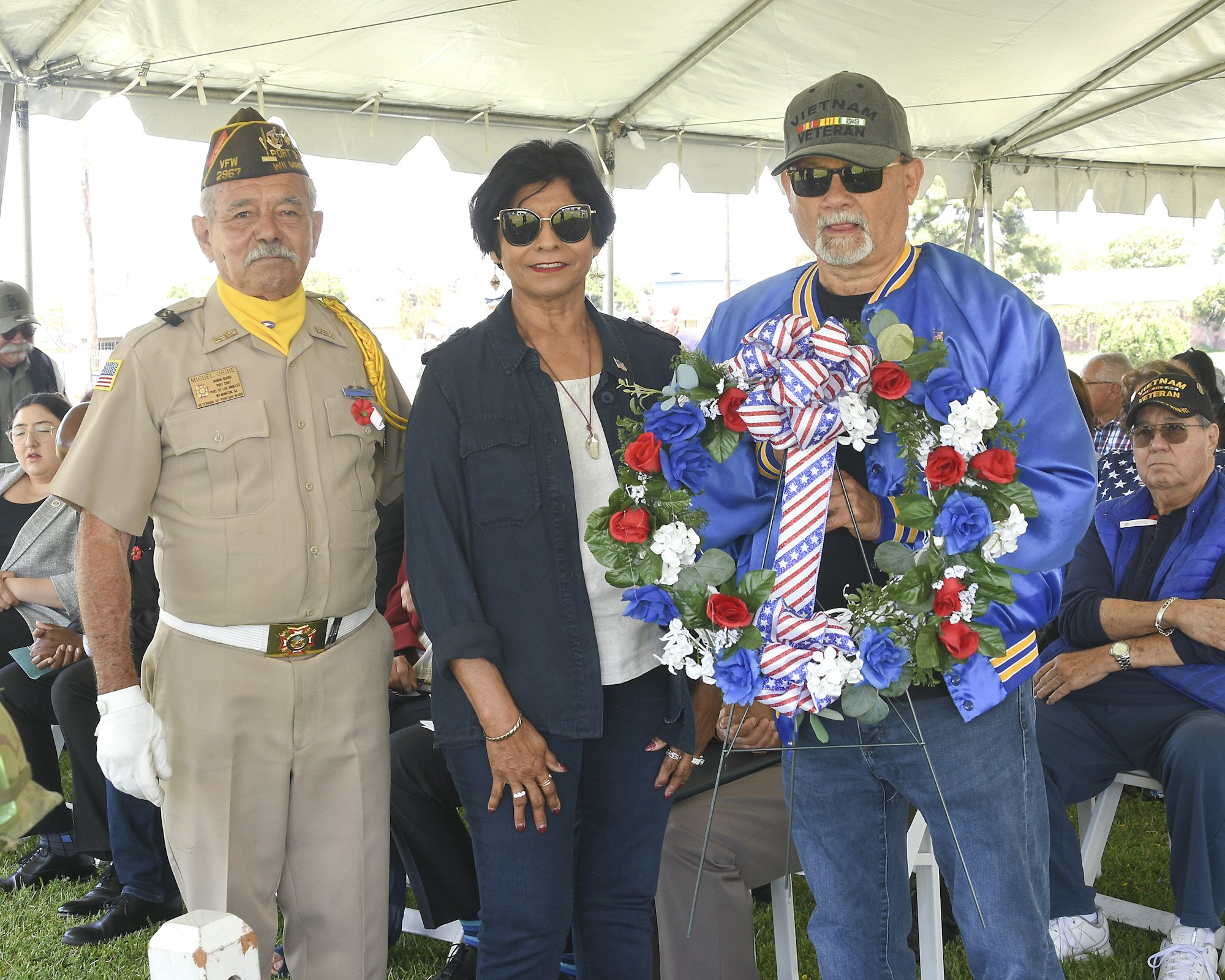 Three people at an event, one in military regalia, another holding a floral wreath, possibly a commemorative gathering.