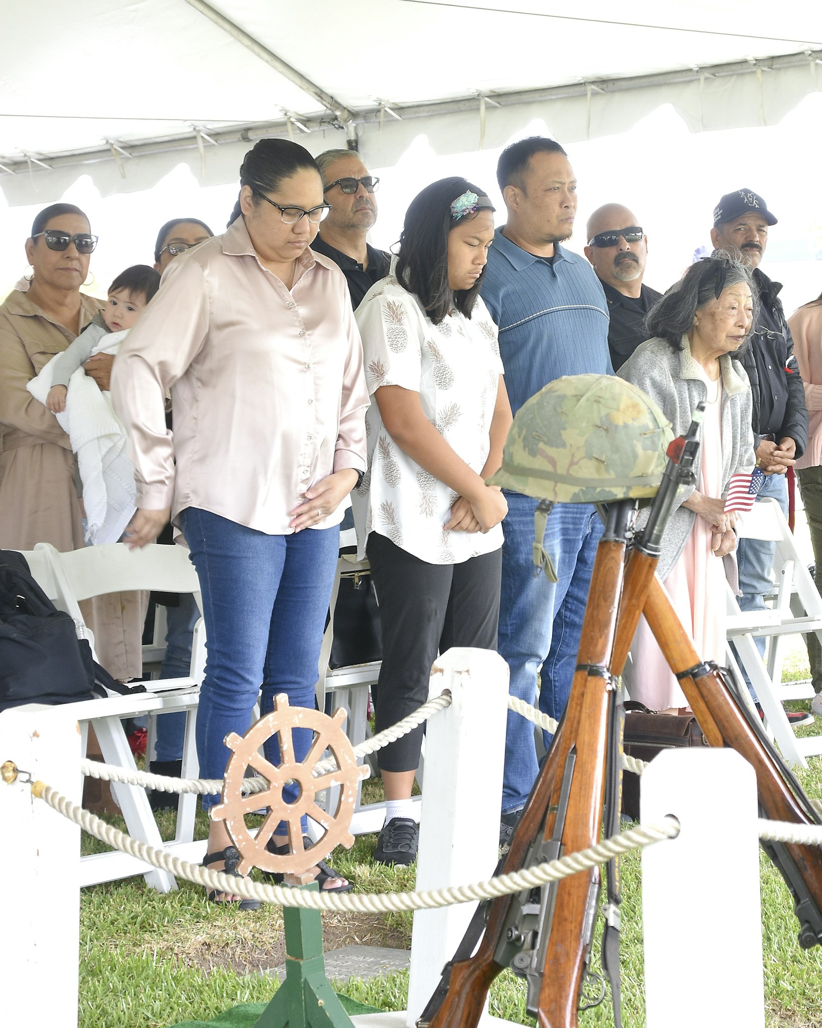 A group of people observing something, standing behind a rope barrier with visible military helmets and rifles on display.