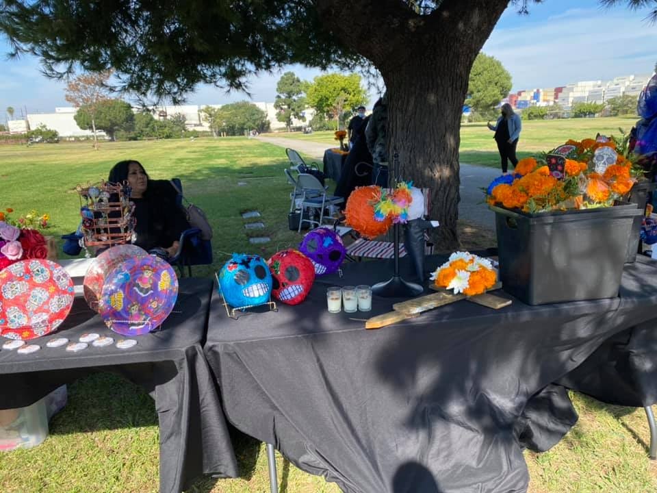 A table displays colorful art for a celebration, featuring decorated skulls, candles, and flowers, with people enjoying the event.