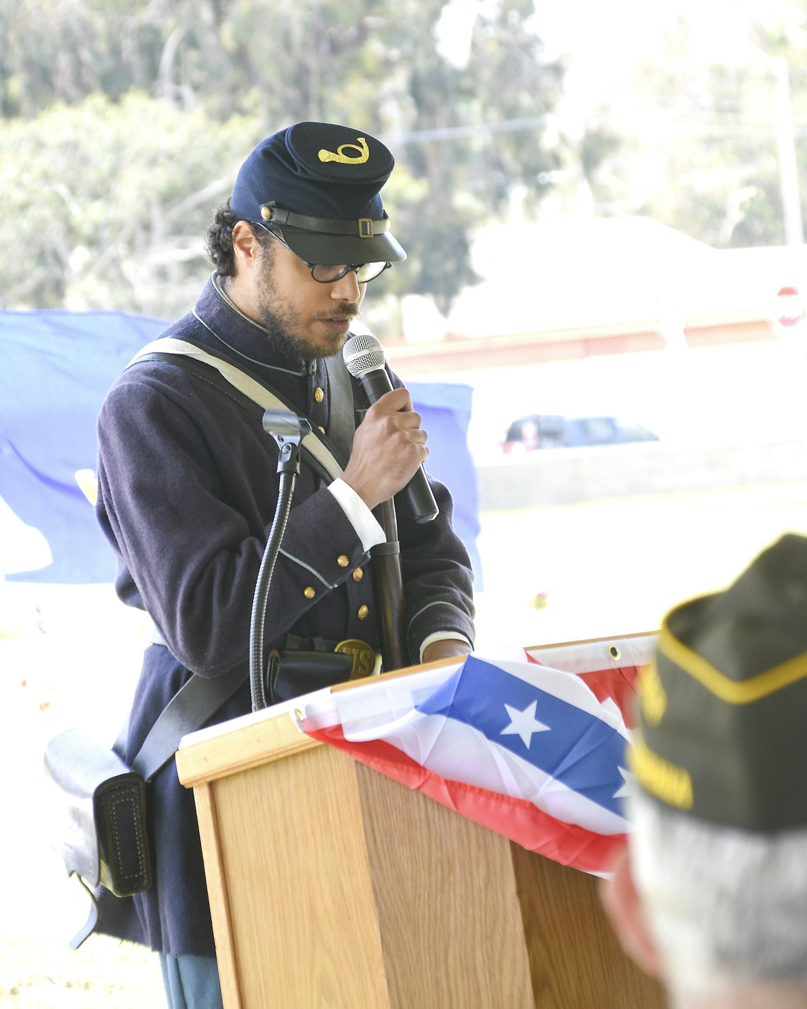 Person in historical military uniform speaking into a microphone behind a podium with a flag.