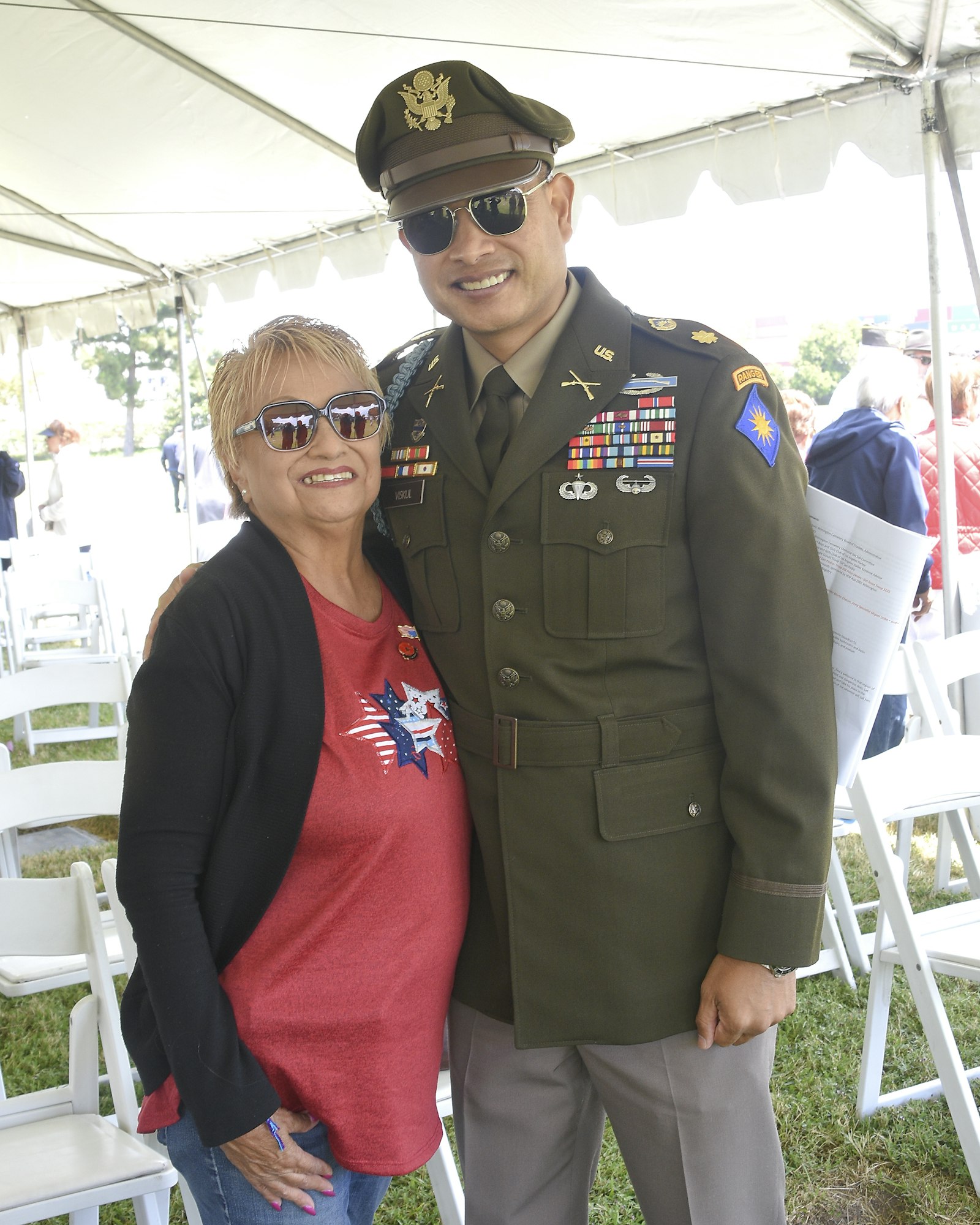 A woman and a military officer in uniform with medals, both smiling, under a white tent.