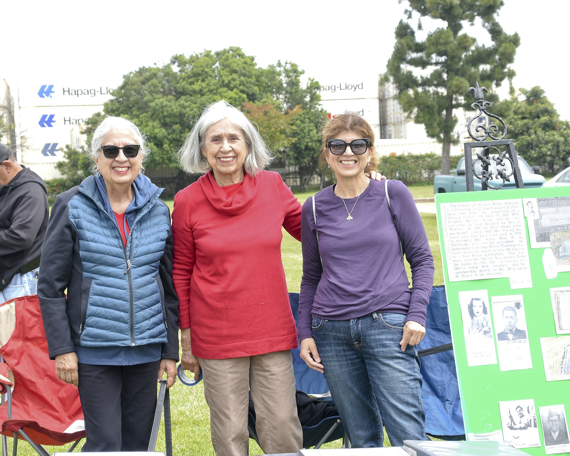 Three smiling women standing together outdoors, behind them a poster with historical information and photos.