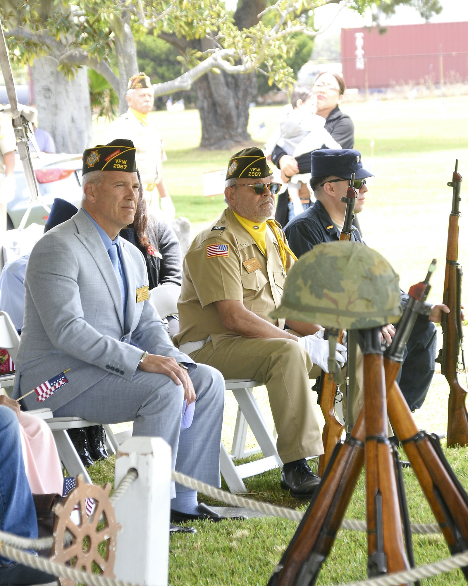 Veterans in uniforms with medals sit at an outdoor event, rifles and a helmet displayed in the foreground.