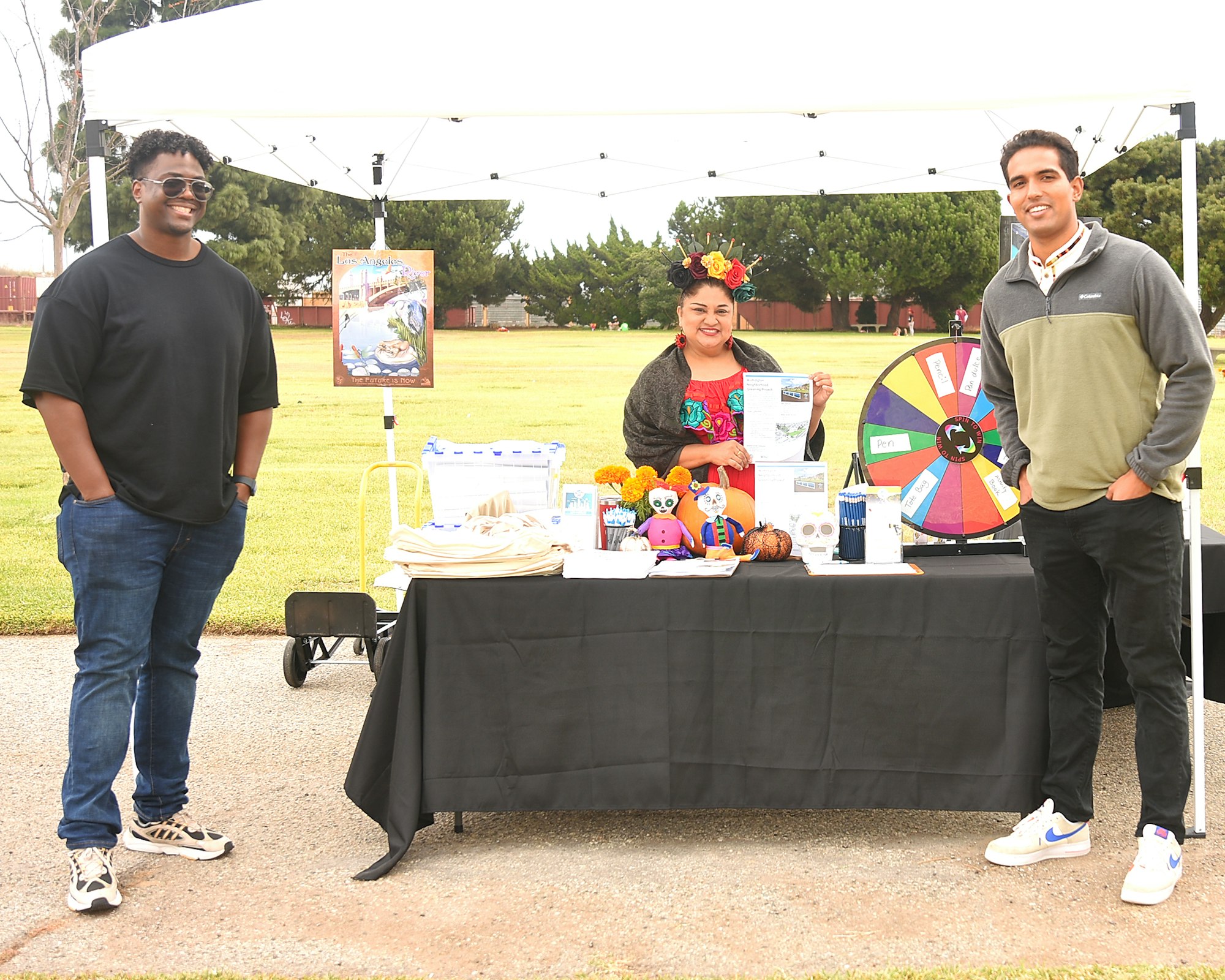 Three people stand by a stall with a prize wheel, flyers, and giveaways on a grassy field.