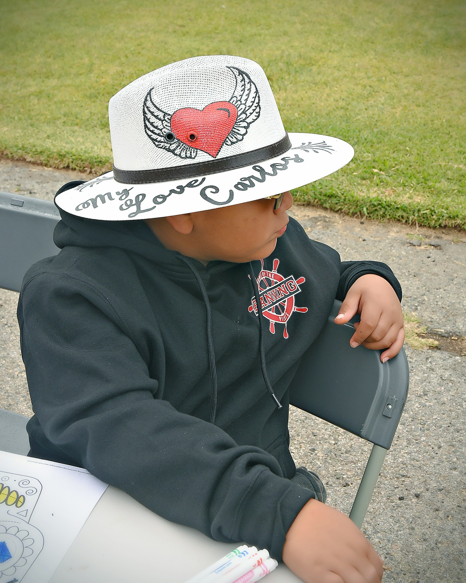 A child with a stylish hat featuring a winged heart, sitting at a table outdoors.