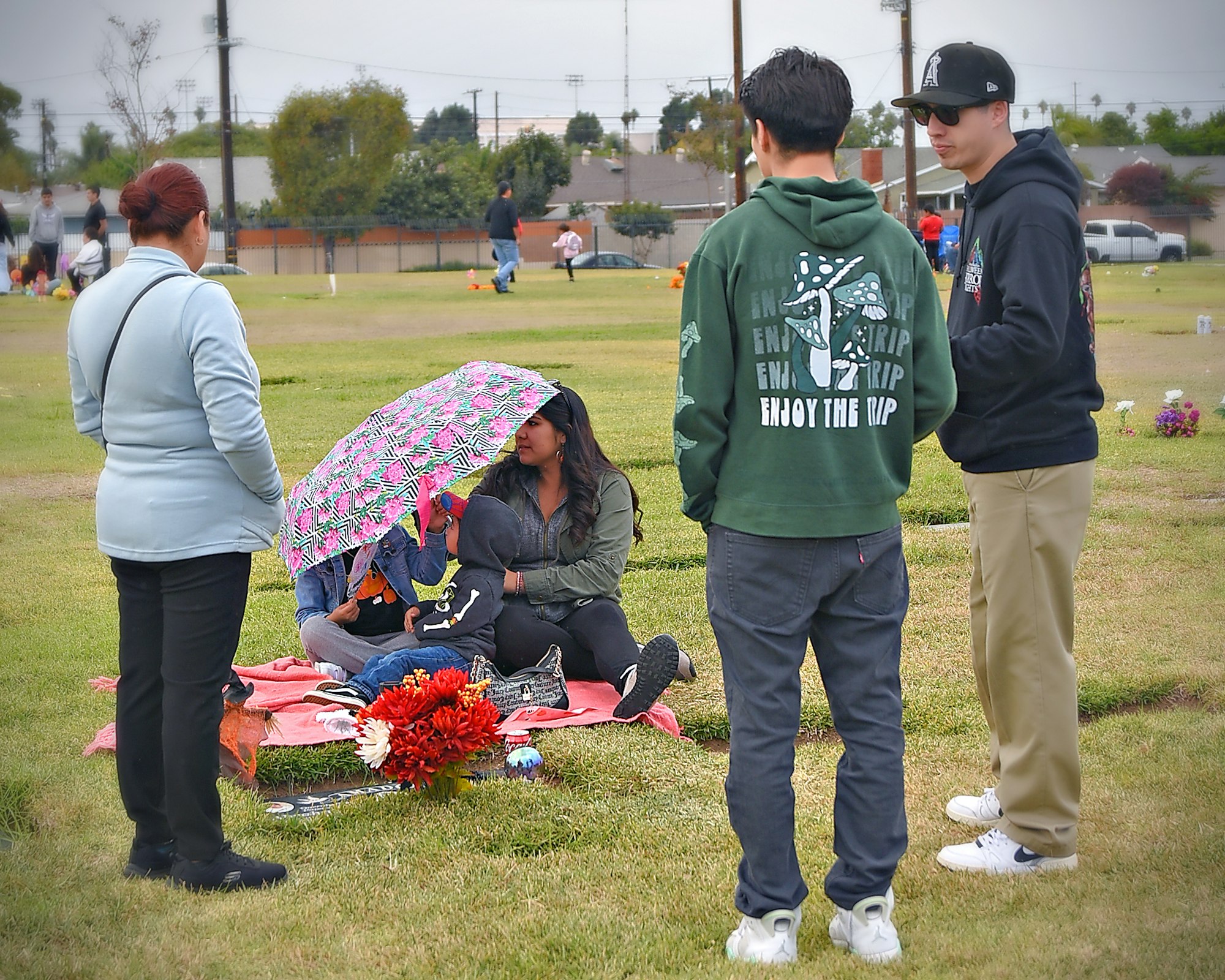 Five people gathered in a park; two standing, a seated group under a pink umbrella, flowers on the ground, and a grassy field in the background.