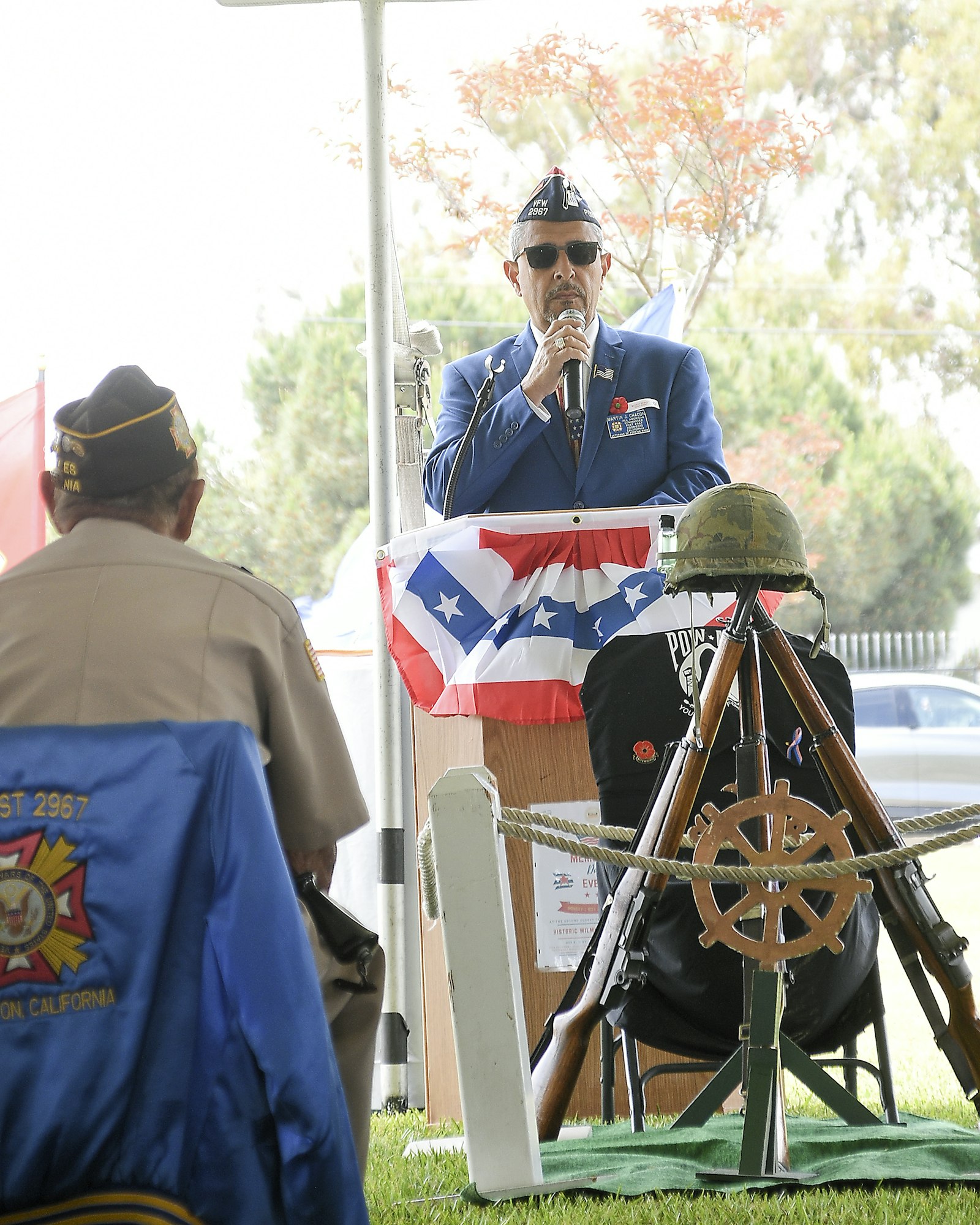 Man speaking into microphone at a veteran event, another listening, with US flag decor and a military helmet.