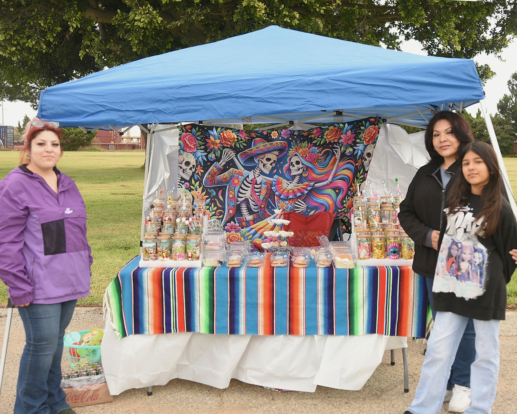 A stall with colorful textiles, snacks, and a Day of the Dead themed backdrop, attended by two adults and a child.